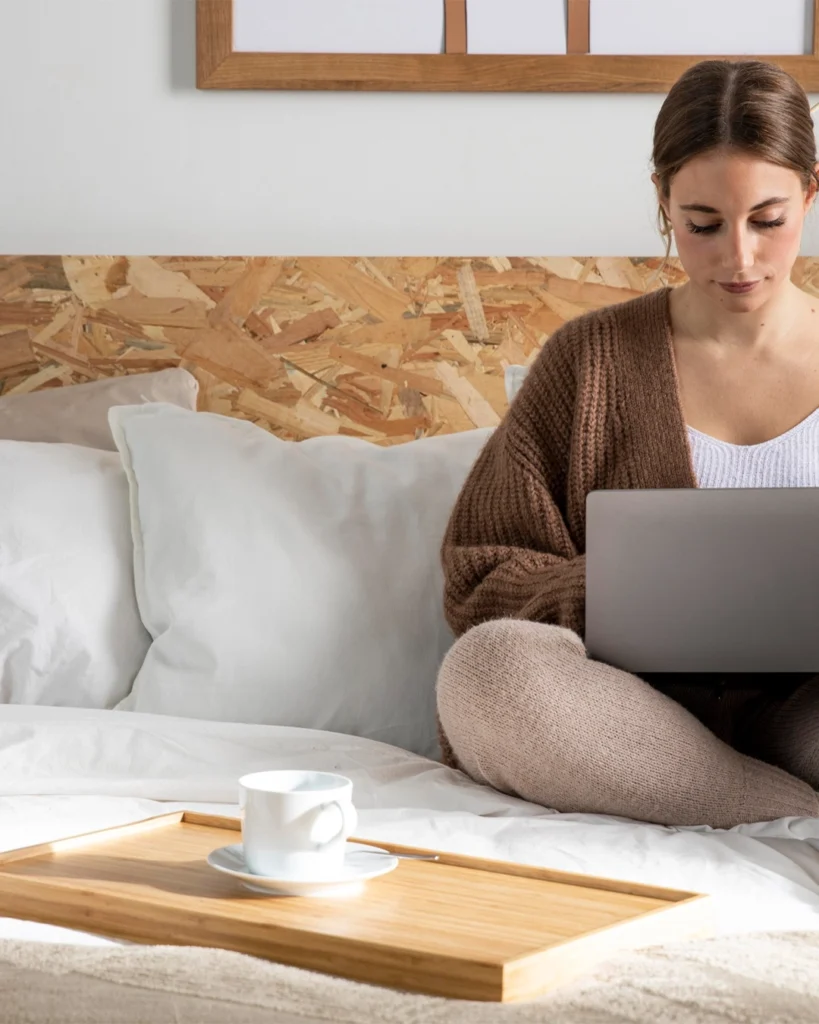 Žena s počítačom na kolenách sedí na posteli, vedľa seba má šálku s kávou a oddychuje